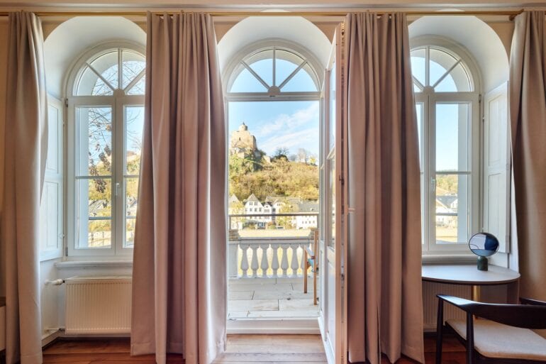Zwei hohe Fenster und eine mittige Tür trennen ein Altbauzimmer mit Parkettboden von einem weoßen Balkon. Der Blick geht auf eine Burgruine in einem Wald.