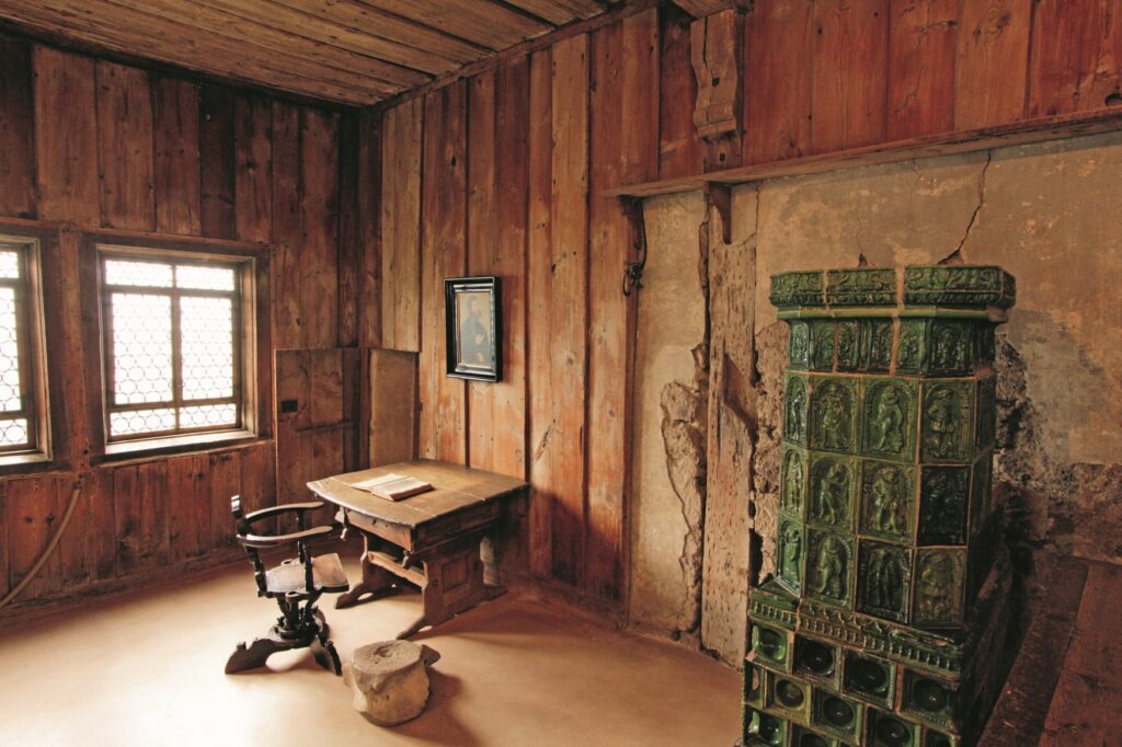 A wood-paneled room in a castle. On the left are two glass windows, on the right wall is a simple wooden table with a chair and a book, and to the right of that is a green tiled stove.