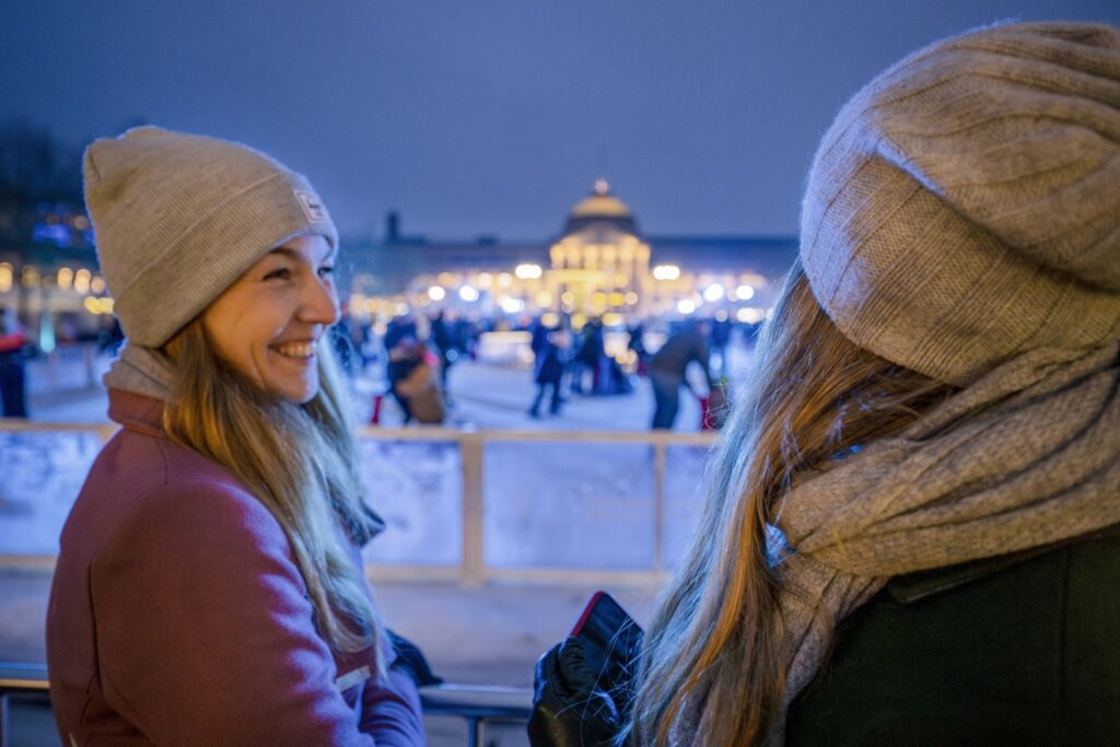 Two women are standing at the edge of an ice rink in a large square, both wearing hats. The woman on the left is smiling at the other woman, whose face cannot be seen. The Wiesbaden State Theater can be seen blurred in the background, and many people are skating on the ice rink itself.