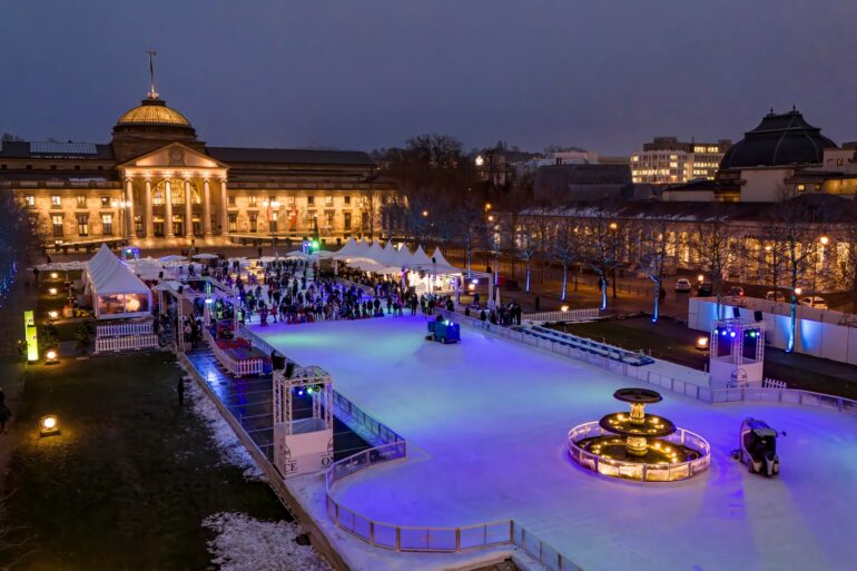 In front of the classicist State Theater in Wiesbaden, brightly lit, there is a long ice rink where many people are skating.