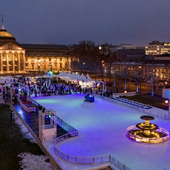 Vor dem klassizistischen Staatstheater Wiesbaden, hell erleuchtet, liegt eine lange Eisbahn, auf der viele Menschen fahren.