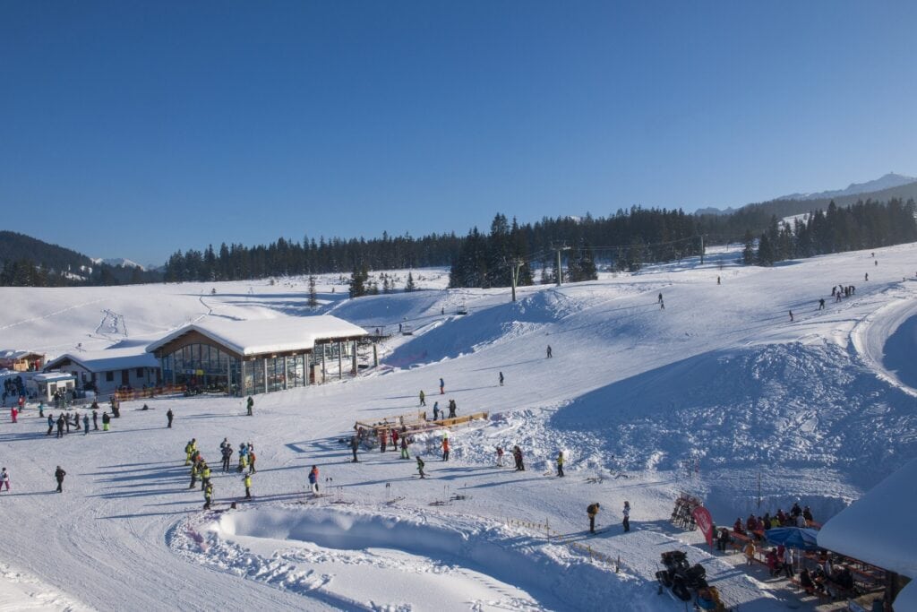 Der Auslauf einer Skipiste in den Alpen, auf der viele Menschen unterwegs sind. Links der Einstieg zum Skilift, rechts die Piste, die langsam zum linken Bildrand ausläuft. Dahinter Bäume und blauer Himmel.