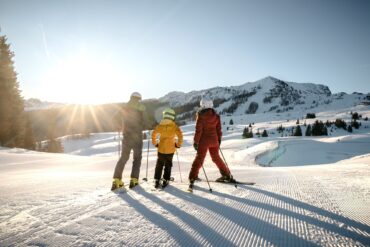 Drei Personen in Winterkleidung stehen mit dem Rücken zum Betrachter auf Skiern auf einer Piste und blicken in Richtung einer dicht verschneiten Bergkette. Vom Himmel strahlt hell die Sonne.