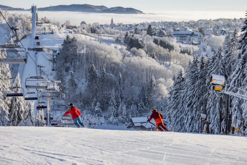Two people are skiing down a slope. To their left is a ski lift, and to their right, in the background, on the other side of the valley, is a small village.