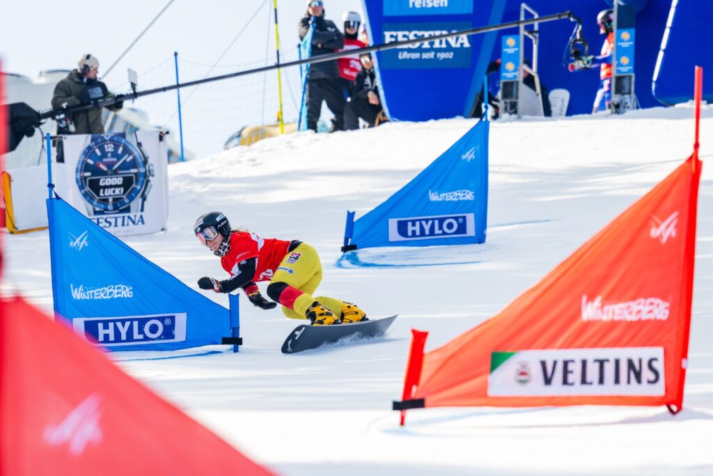 A woman in colorful winter sports clothing, wearing a helmet and goggles, slaloms between several flags on a snowboard. The flags feature advertising prints and the word “Winterberg.”