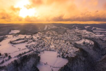 Aerial view of a small town in a low mountain range. Everything is covered in snow, and dense coniferous forest surrounds the town. In the sky, the sun sets behind the clouds, bathing everything in a golden yellow light.