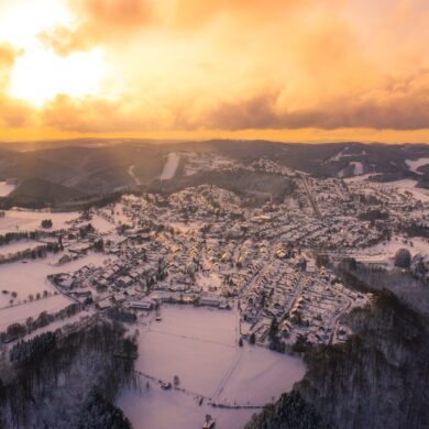 Luftaufnahme einer Kleinstadt in einem Mittelgebirge. Alles ist von Schnee bedeckt, rund um den Ort erstreckt sich dichter Nadelwald. Am Himmel geht die Sonne hinter den Wolken unter und taucht alles in ein goldgelbes Licht.