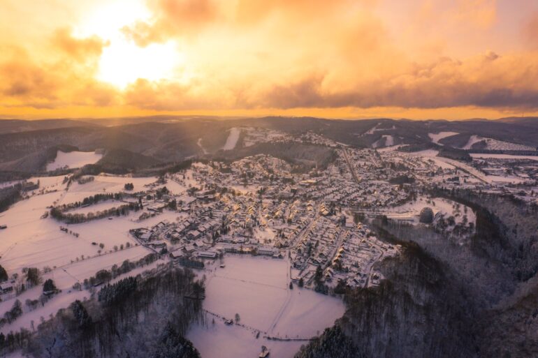 Luftaufnahme einer Kleinstadt in einem Mittelgebirge. Alles ist von Schnee bedeckt, rund um den Ort erstreckt sich dichter Nadelwald. Am Himmel geht die Sonne hinter den Wolken unter und taucht alles in ein goldgelbes Licht.
