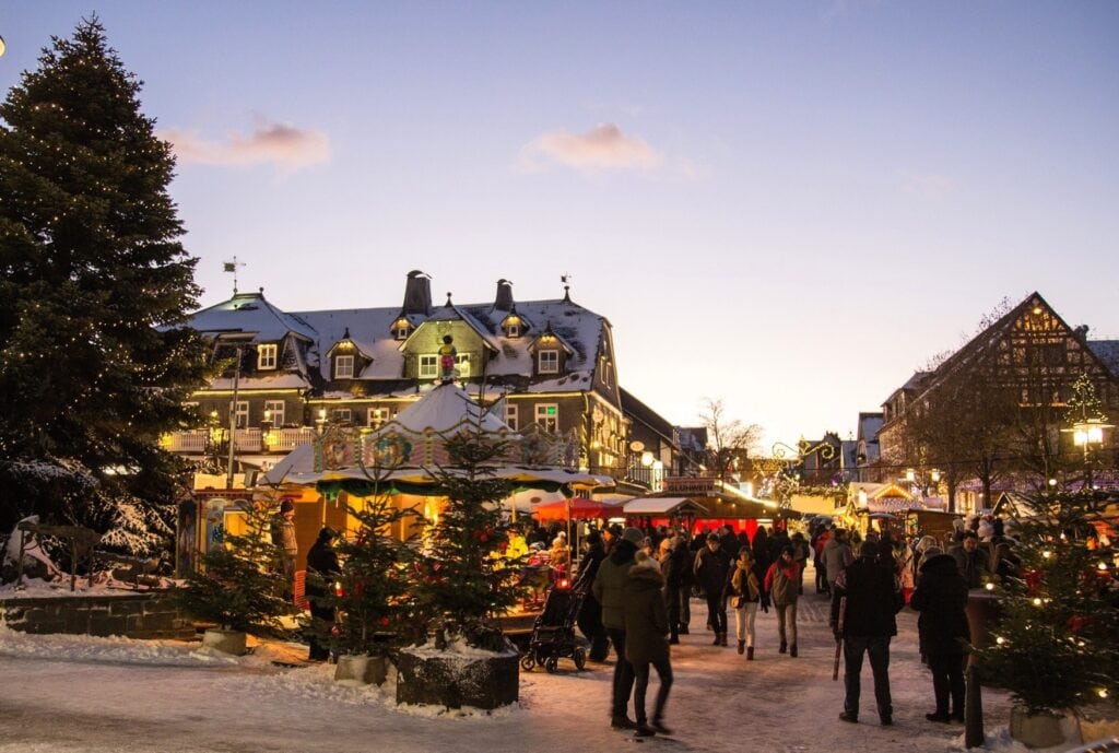 Auf einem verschneiten Marktplatz, umgeben von beleuchteten Fachwerkhäusern, sind viele Menschen auf einem Weihnachtsmarkt unterwegs. Am Himmel dämmert es bereits.