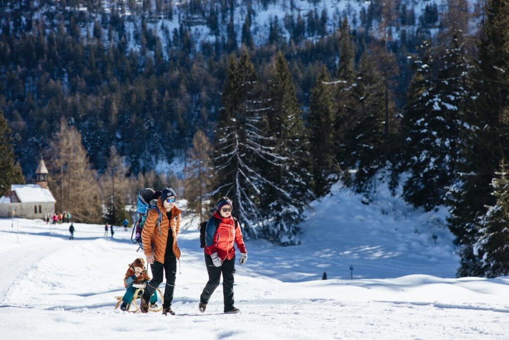 Zwei junge Eltern in Winterkleidung ziehen ihr Kind auf einem Holzschlitten hinter sich her, während sie auf einem Winterwanderpfad durch den Schnee hinterlaufen. Im Hintergrund ein Berghang mit dichtem Wald.