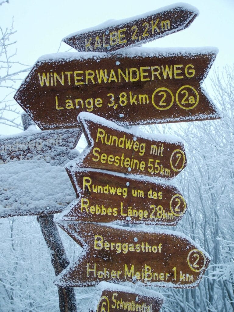 Ein Wegweiser mit mehreren Schildern daran, alle von feinem Pulverschnee bedeckt. Das größte Schild ganz oben zeigt den "Winterwanderweg" mit einer Länge von 3,8 km.