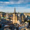 Stadtbild von Aachen mit dem Dom im Zentrum. Am prominentesten eine achteckige Kuppel in der Mitte, rechts daneben der Kirchenchor mit hohen gotischen Fenster.