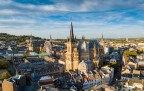 Stadtbild von Aachen mit dem Dom im Zentrum. Am prominentesten eine achteckige Kuppel in der Mitte, rechts daneben der Kirchenchor mit hohen gotischen Fenster.