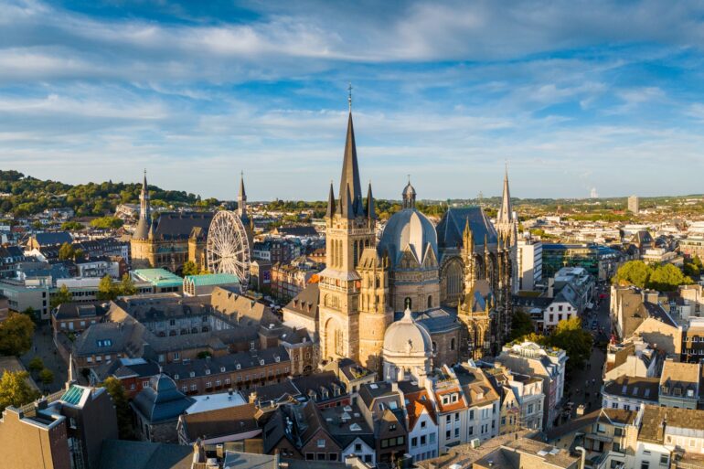 Stadtbild von Aachen mit dem Dom im Zentrum. Am prominentesten eine achteckige Kuppel in der Mitte, rechts daneben der Kirchenchor mit hohen gotischen Fenster.