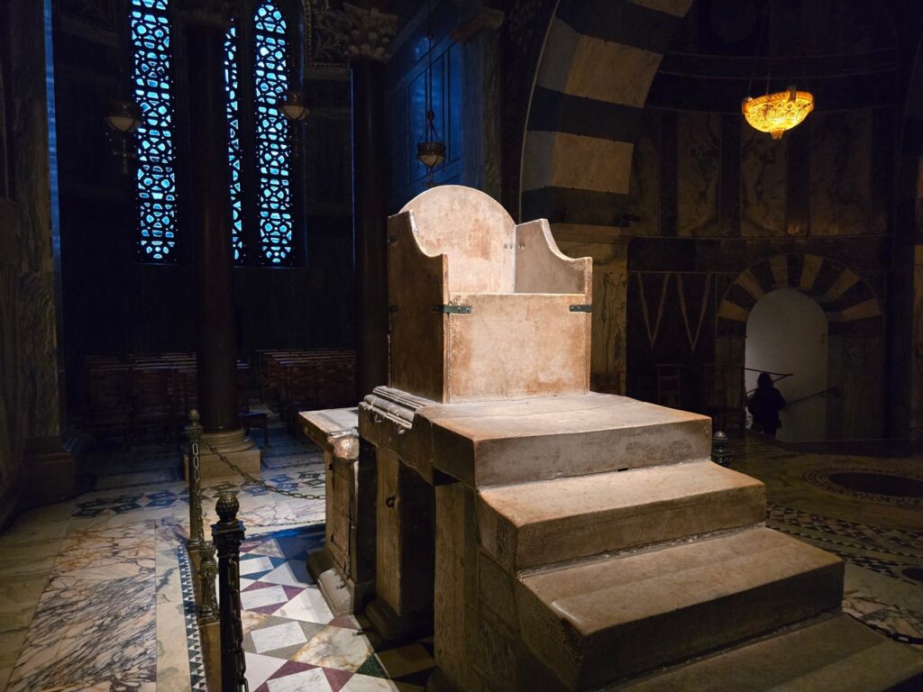 Ein simpler, steinerner Thron steht auf einem Podest mit vier Treppenstufen. Die Fliesen rundherum sind kunstvoll gearbeitet, an der Wand hinter dem Thron fällt etwas Licht durch zwei längliche Buntfenster.