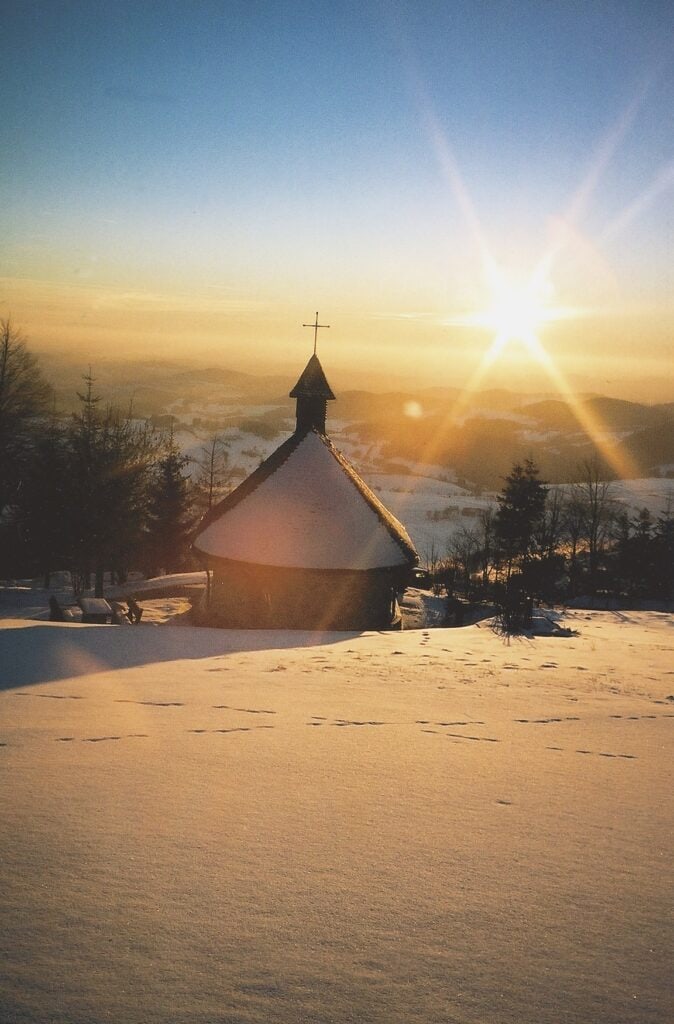Auf einer Anhöhe, beschienen von der Sonne und umgeben von frischem Schnee, steht eine kleine, halbrunde Wallfahrtskapelle mit einem Kreuz auf dem Türmchen. Dahinter öffnet sich ein verschneites Tal mit vielen Waldstücken.