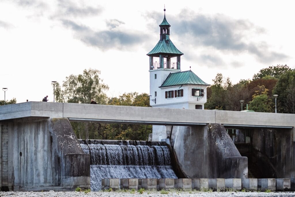 Von links nach rechts zieht sich der Ablass eines Beton-Staudamms durchs Bild. Mittig aufgesetzt it ein kleines, weißes Gebäude mit einem eckigen Turm, beide mit Messingdach.