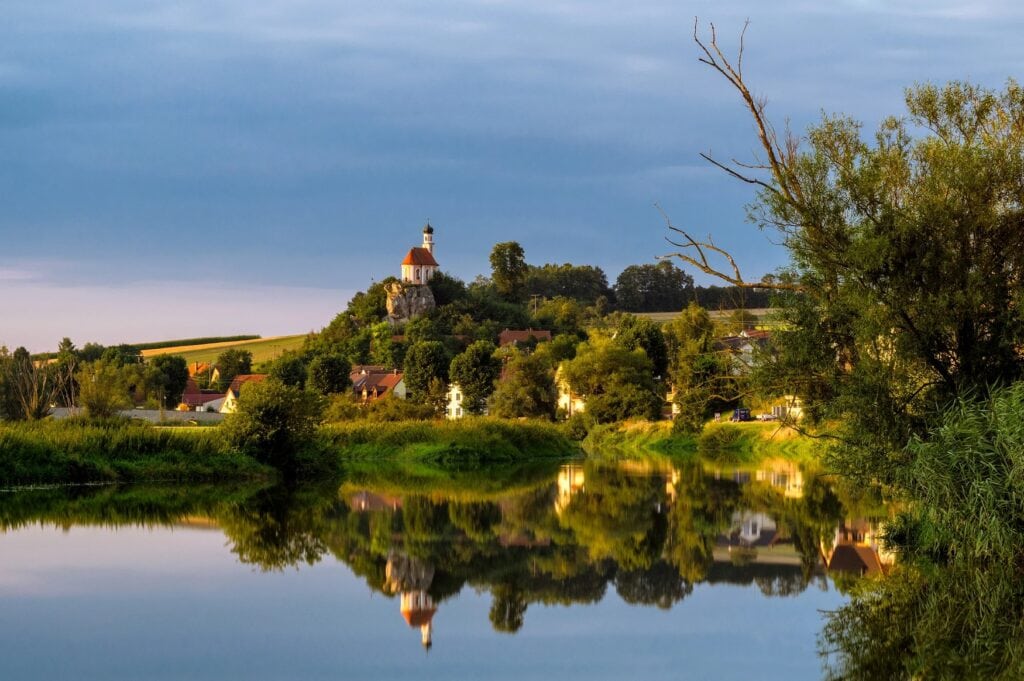 Um das Ufer eines Sees herum liegen mehrere weiße Häuser mit roten Dächern. Ein hoher Felsen ragt ein Stück in den See, darauf steht eine kleine Kapelle. Alles spiegelt sich in der stillen Wasseroberfläche.