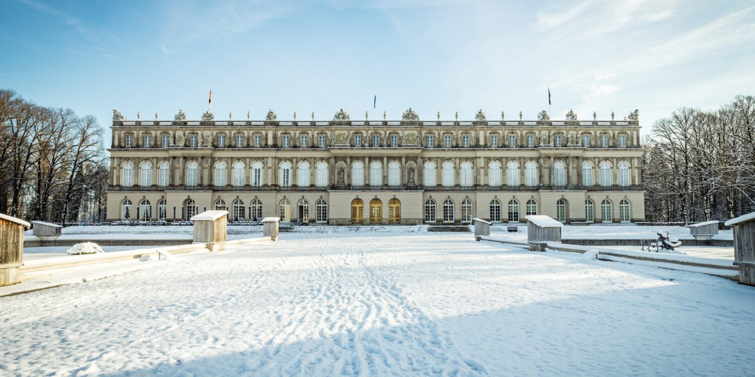 Hinter einer zugeschneiten Zufahrt liegt ein breites, weißes, dreigeschossiges Barockschloss. Darüber blauer Himmel.