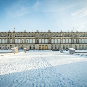 Hinter einer zugeschneiten Zufahrt liegt ein breites, weißes, dreigeschossiges Barockschloss. Darüber blauer Himmel.