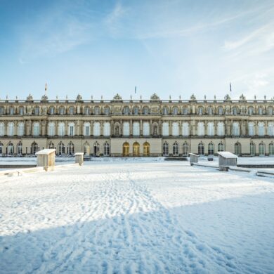 Hinter einer zugeschneiten Zufahrt liegt ein breites, weißes, dreigeschossiges Barockschloss. Darüber blauer Himmel.
