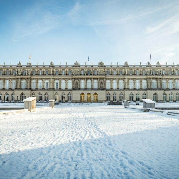 Hinter einer zugeschneiten Zufahrt liegt ein breites, weißes, dreigeschossiges Barockschloss. Darüber blauer Himmel.