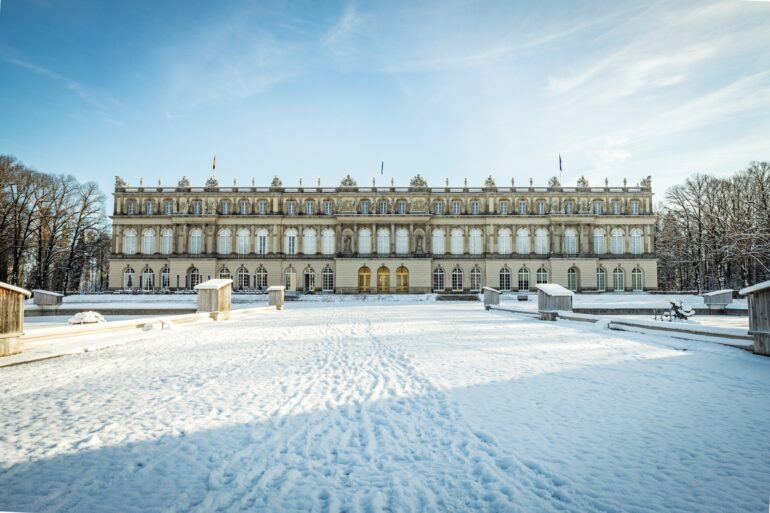 Hinter einer zugeschneiten Zufahrt liegt ein breites, weißes, dreigeschossiges Barockschloss. Darüber blauer Himmel.