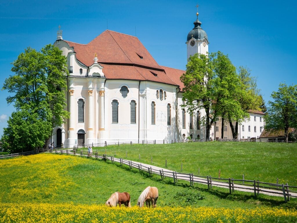 Hinter einer sachten, grünen Anhöhe ragt eine weiße Rokoko-Kirche mit ziegelrotem Dach auf.