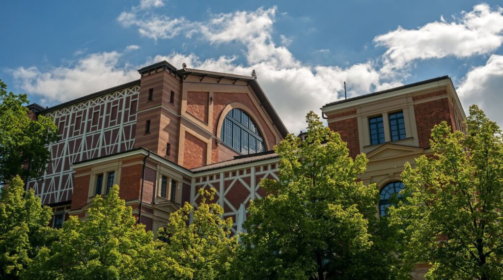 A historic brick building with white half-timbered beams rises out of a green forest. Above it, blue sky.