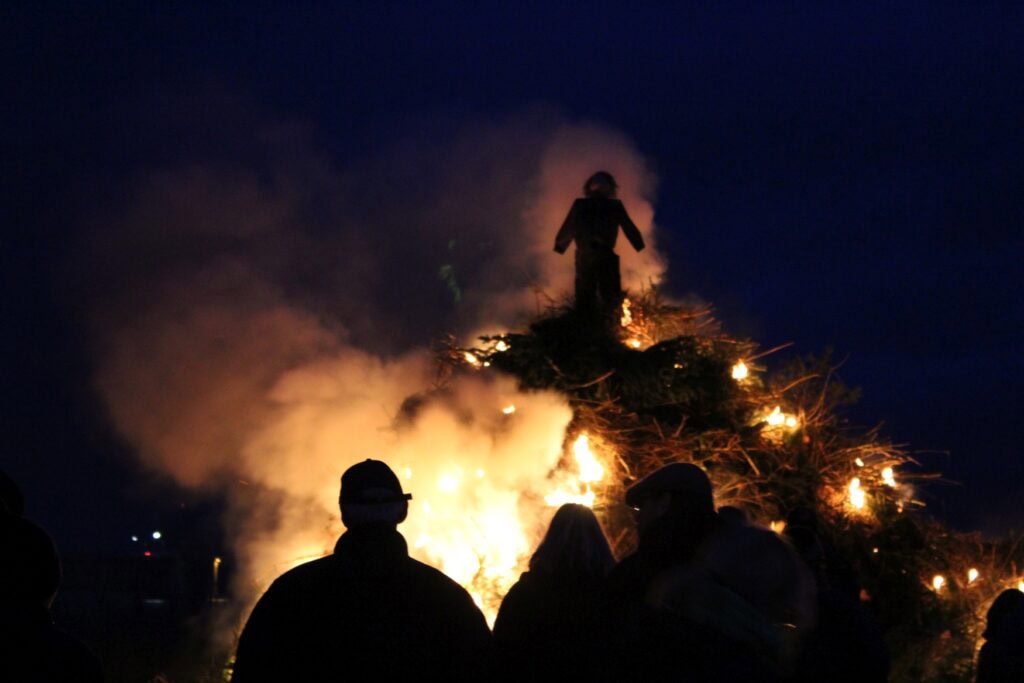 Auf einem Feuerhaufen brennt eine Strohpuppe vor dem Nachthimmel. Im Vordergrund stehen einige Personen, die nur als Silhouetten erkennbar sind.