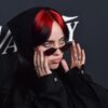 Nahaufnahme von Billie Eilish mit roten Haaren und einer schmalen Sonnenbrille, im schwarzen Hoodie, vor einem dunklen Backdrop.