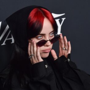 Nahaufnahme von Billie Eilish mit roten Haaren und einer schmalen Sonnenbrille, im schwarzen Hoodie, vor einem dunklen Backdrop.
