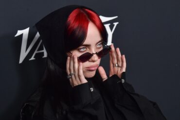 Nahaufnahme von Billie Eilish mit roten Haaren und einer schmalen Sonnenbrille, im schwarzen Hoodie, vor einem dunklen Backdrop.