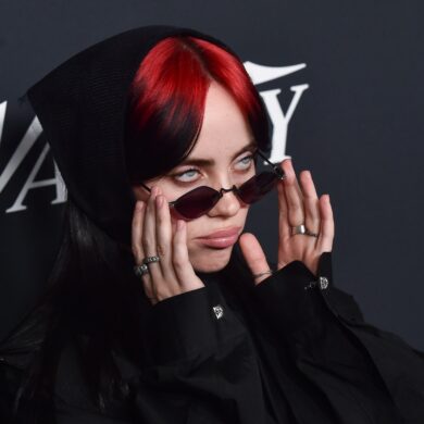 Nahaufnahme von Billie Eilish mit roten Haaren und einer schmalen Sonnenbrille, im schwarzen Hoodie, vor einem dunklen Backdrop.