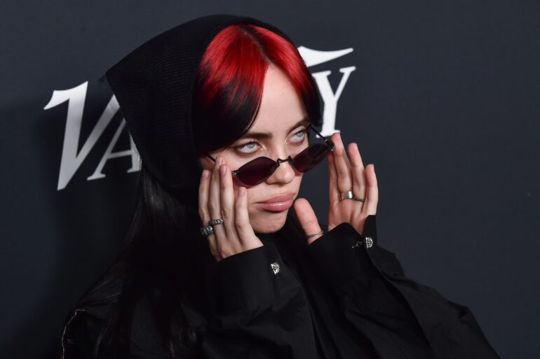 Nahaufnahme von Billie Eilish mit roten Haaren und einer schmalen Sonnenbrille, im schwarzen Hoodie, vor einem dunklen Backdrop.