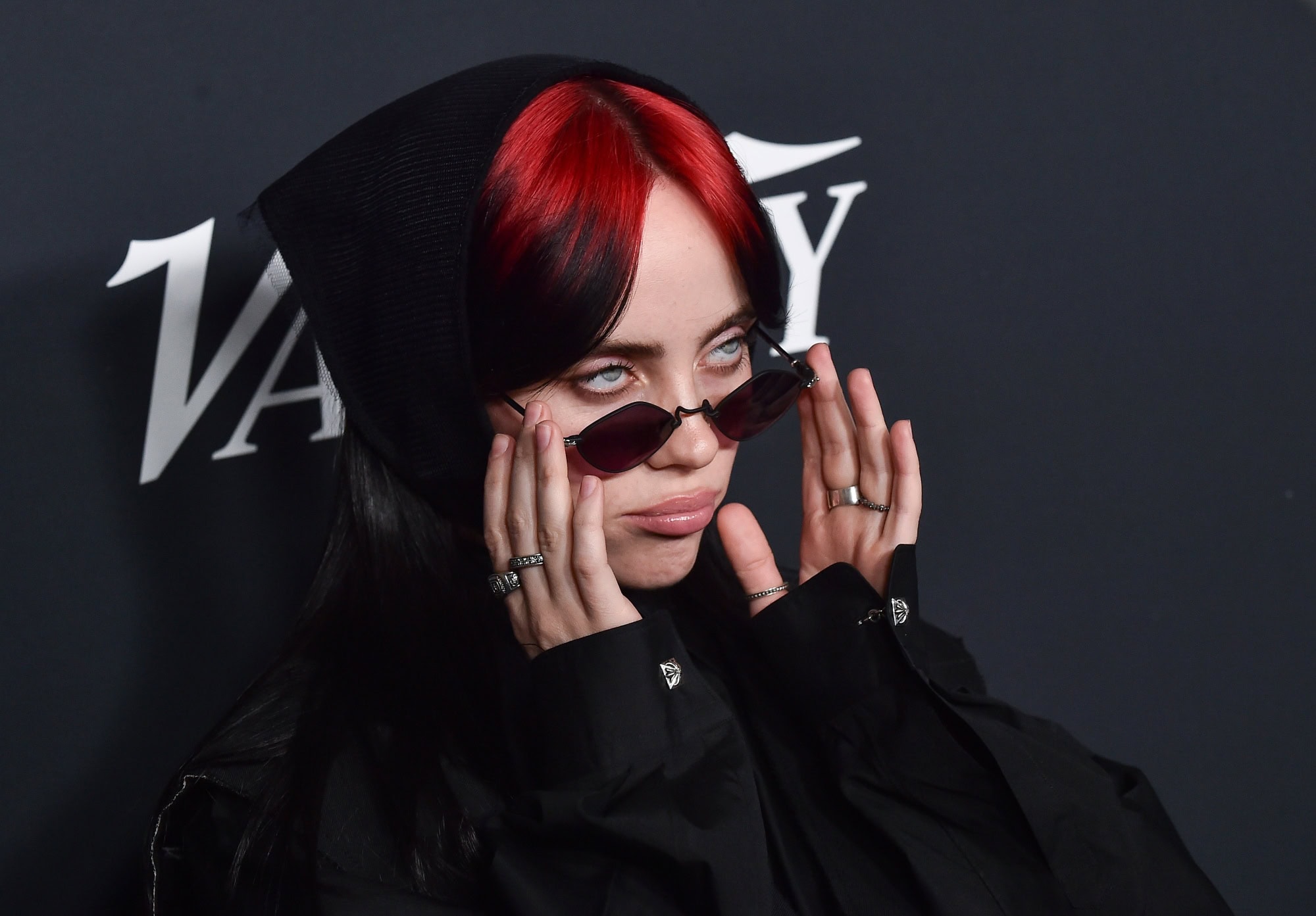 Nahaufnahme von Billie Eilish mit roten Haaren und einer schmalen Sonnenbrille, im schwarzen Hoodie, vor einem dunklen Backdrop.