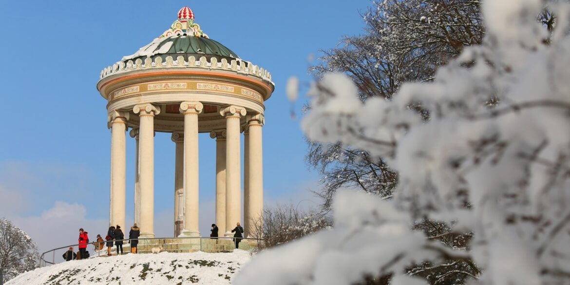 Auf einer verschneiten Anhöhe steht der kreisrunde Monopteros. Viele Menschen sind davor im Park unterwegs.
