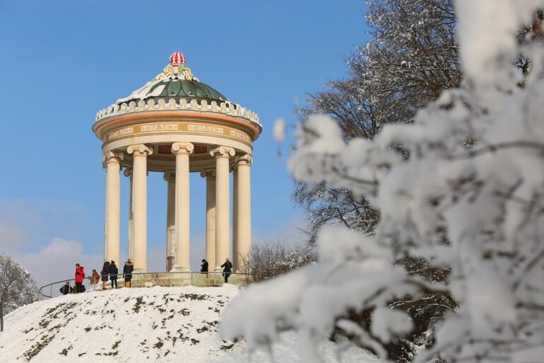 The circular Monopteros stands on a snow-covered hill. Many people are walking in the park in front of it.