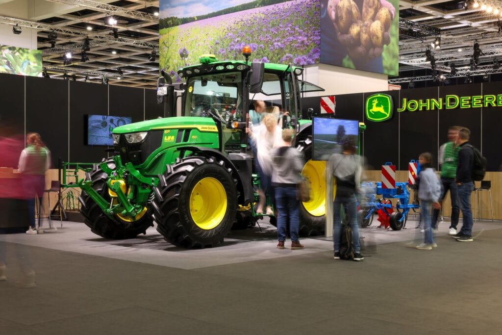 An einem Messestand steht ein großer, grüner Traktor der Marke "John Deere". Einige Personen steigen gerade aus, durch eine Langzeitbelichtung sind sie nur verschwommen zu sehen.