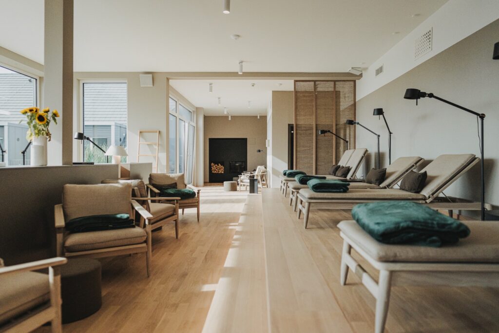 Relaxation room in a hotel spa, lots of wood and white walls; on the right, a raised area with four reclining chairs with gray upholstery lined up in a row; on the left, two gray upholstered armchairs each forming a seating area. Further in the background is an area with a modern fireplace and firewood; daylight shines into the room through a glass front on the left.
