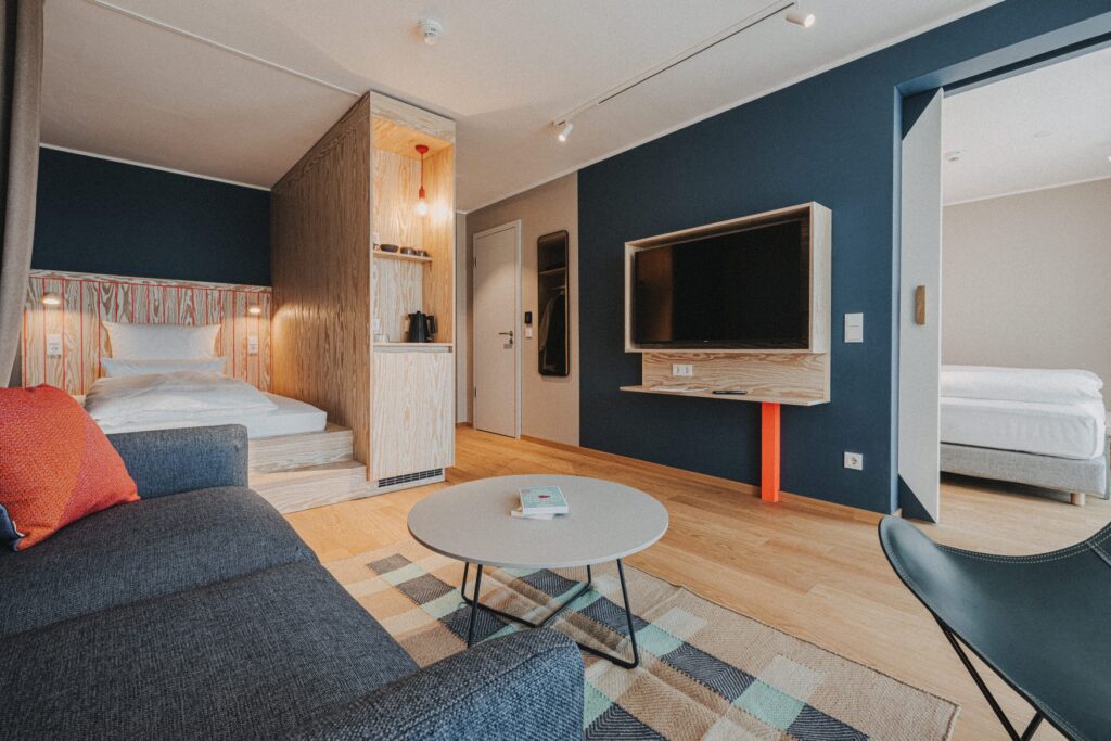 A modern, simple hotel room with blue walls and lots of light wood. On the left in the foreground is an upholstered couch with a round table, diagonally behind it is a niche with a platform bed. In the center is the door to the hallway, to the right of it is a large TV. To the right of the TV is an open door leading to another room with a bed.