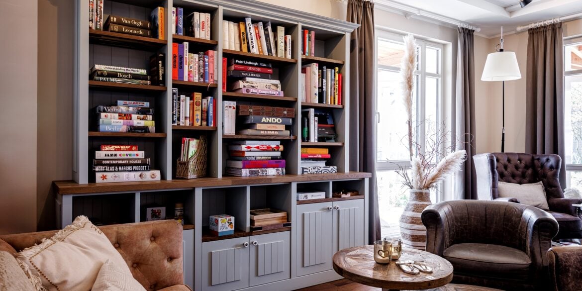 Die Bibliothek im Hotel "Zweite Heimat" in St. Peter-Ording; im Vordergrund eine Ledercouch mit hölzernem Couchtisch und Ledersessel gegenüber, an der Wand dahinter ein graues, vollgepacktes Holzregal. Rechts im Bild an der Ecke je ein bodentiefes Fenster.