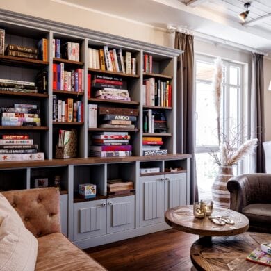 Die Bibliothek im Hotel "Zweite Heimat" in St. Peter-Ording; im Vordergrund eine Ledercouch mit hölzernem Couchtisch und Ledersessel gegenüber, an der Wand dahinter ein graues, vollgepacktes Holzregal. Rechts im Bild an der Ecke je ein bodentiefes Fenster.