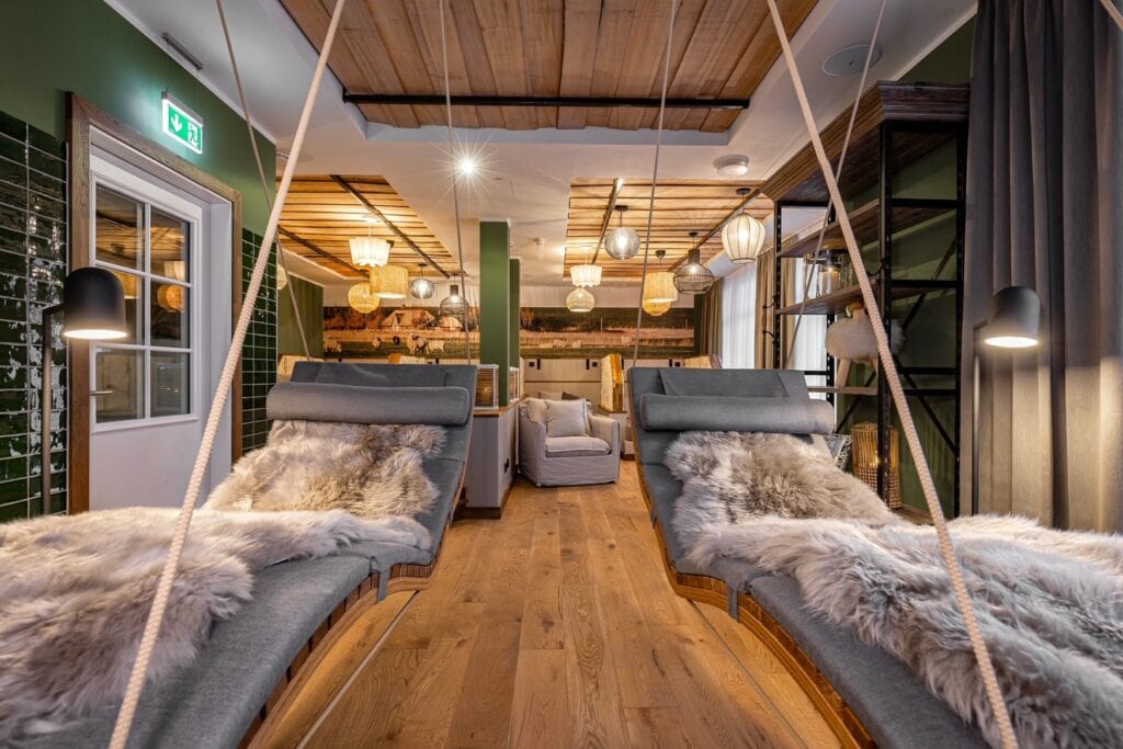 Ruheraum in einem Spa; Im Vordergrund hängen zwei hölzerne Polsterliegen an Seilen von der Decke, dahinter links eine Anrichte und rechts ein Polstersessel auf dem hölzernen Dielenboden. Das Gebälk der Decke ist teils freigelegt, teils weiß verputzt, alles hat einen rustikalen Charme.