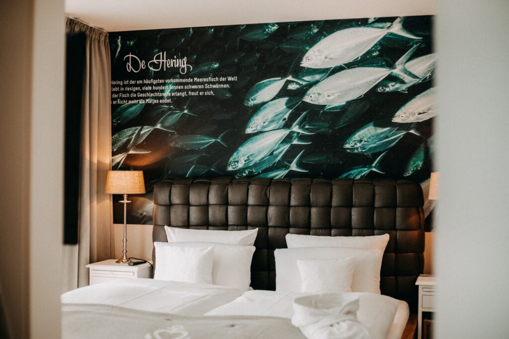 Blick durch eine Tür auf eine breites Doppelbett in einem Hotelzimmer; das Bett ist weiß bezogen, dahinter ein ledernes Kopfteil und darüber ein Wandkunstwerk mit einem Schwarm Heringe und einem Infotext dazu.