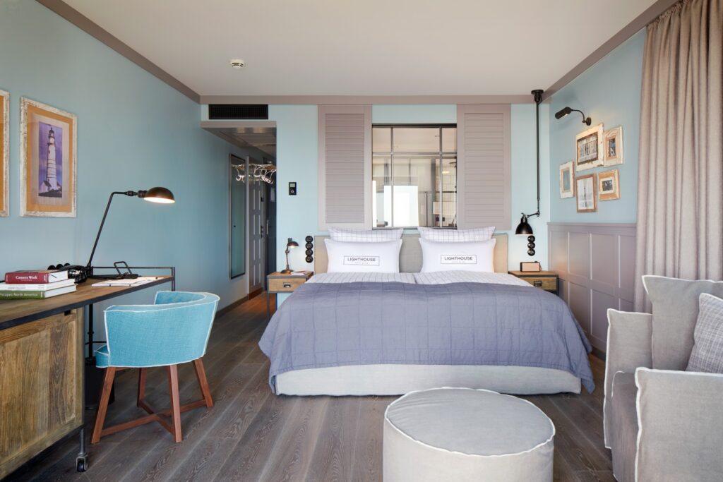 A hotel room, viewed from the window; on the left, a small wooden desk with a blue upholstered chair; in the center, a wide double bed with a bedspread; above it, a window leading to the bathroom. The pillows bear the words “Lighthouse.” The walls are painted a pale turquoise, and several pictures with maritime motifs hang on the walls.