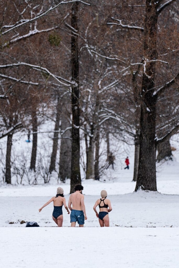 Drei Personen in Schwimmkleidung steigen im verschneiten Englischen Garten in einen Bach.
