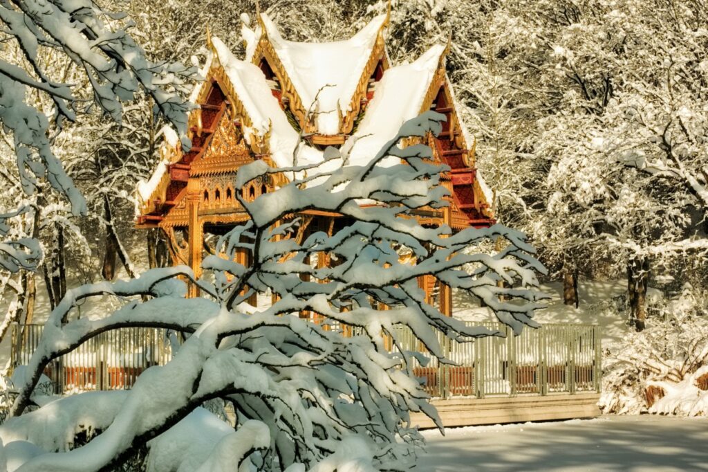A snow-covered, Chinese-style wooden pavilion lies in the middle of a small forest in the winter sun.