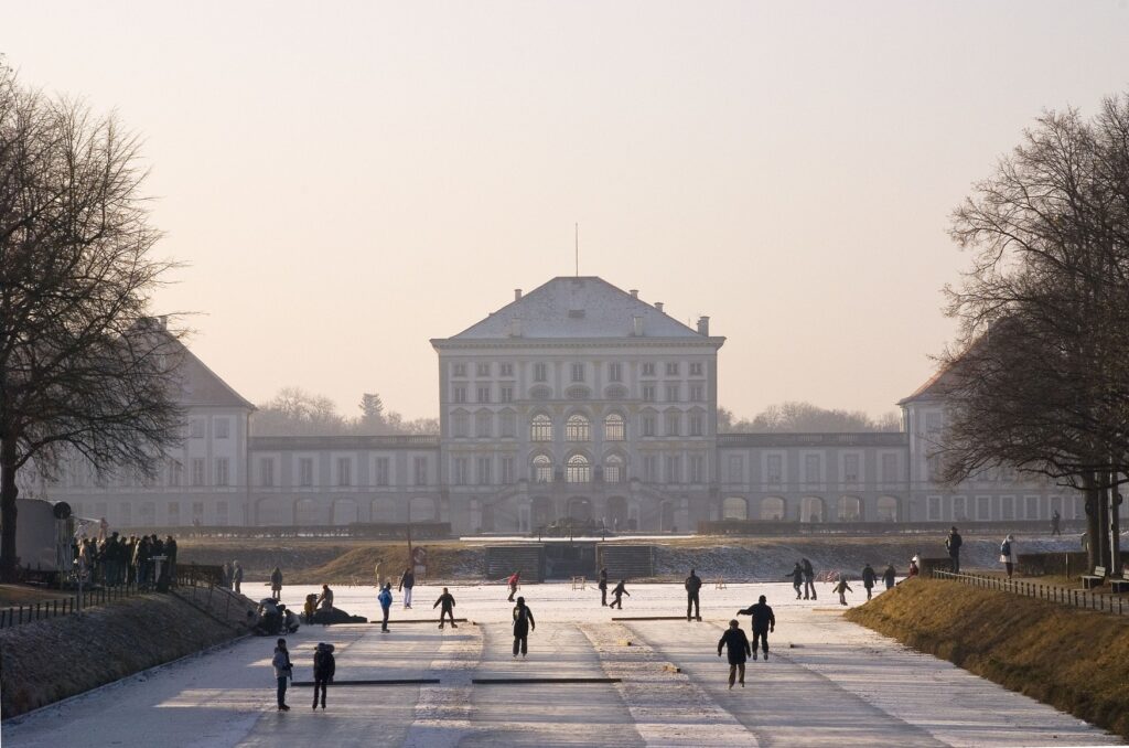 Many people are skating on a long, frozen canal. In the background, the facade of Nymphenburg Palace stretches across the picture from right to left.