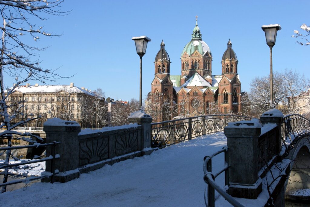 View across a snow-covered bridge over the Isar river to the brick-red St. Luke's Church, with its central brass dome, partially covered in snow.